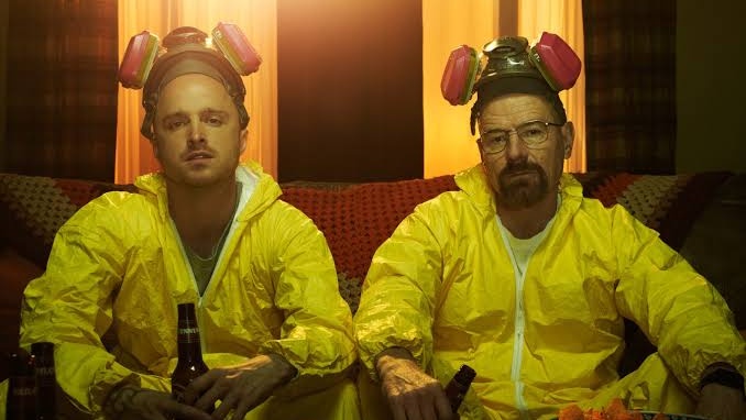 Actors Bryan Cranston and Aaron Paul, wearing yellow hazmat suits, sipping beer on sofa in a scene from Breaking Bad, the hit AMC crime series&nbsp;&ndash; here's where to watch and stream seasons 1-5