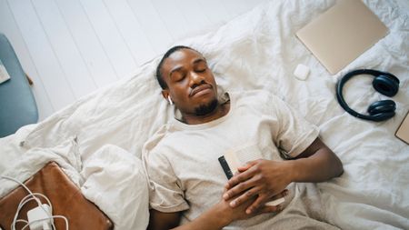 A young man sleeping on a bed while holding a book and listening to his sleep earbuds 