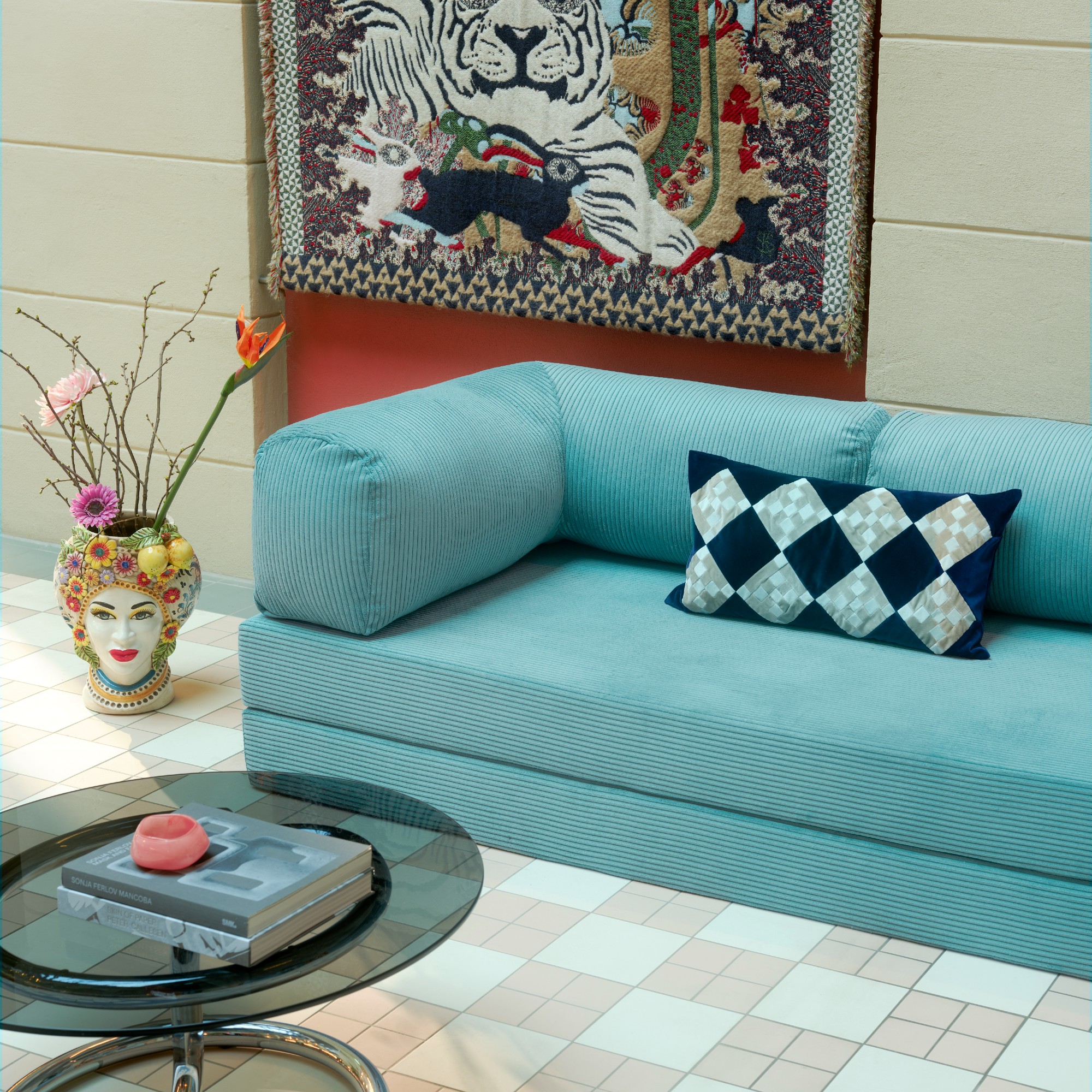The Teddy sofa by OMHU in Turquoise with a head-shaped vase next to it and a round glass and chrome coffee table in front of it