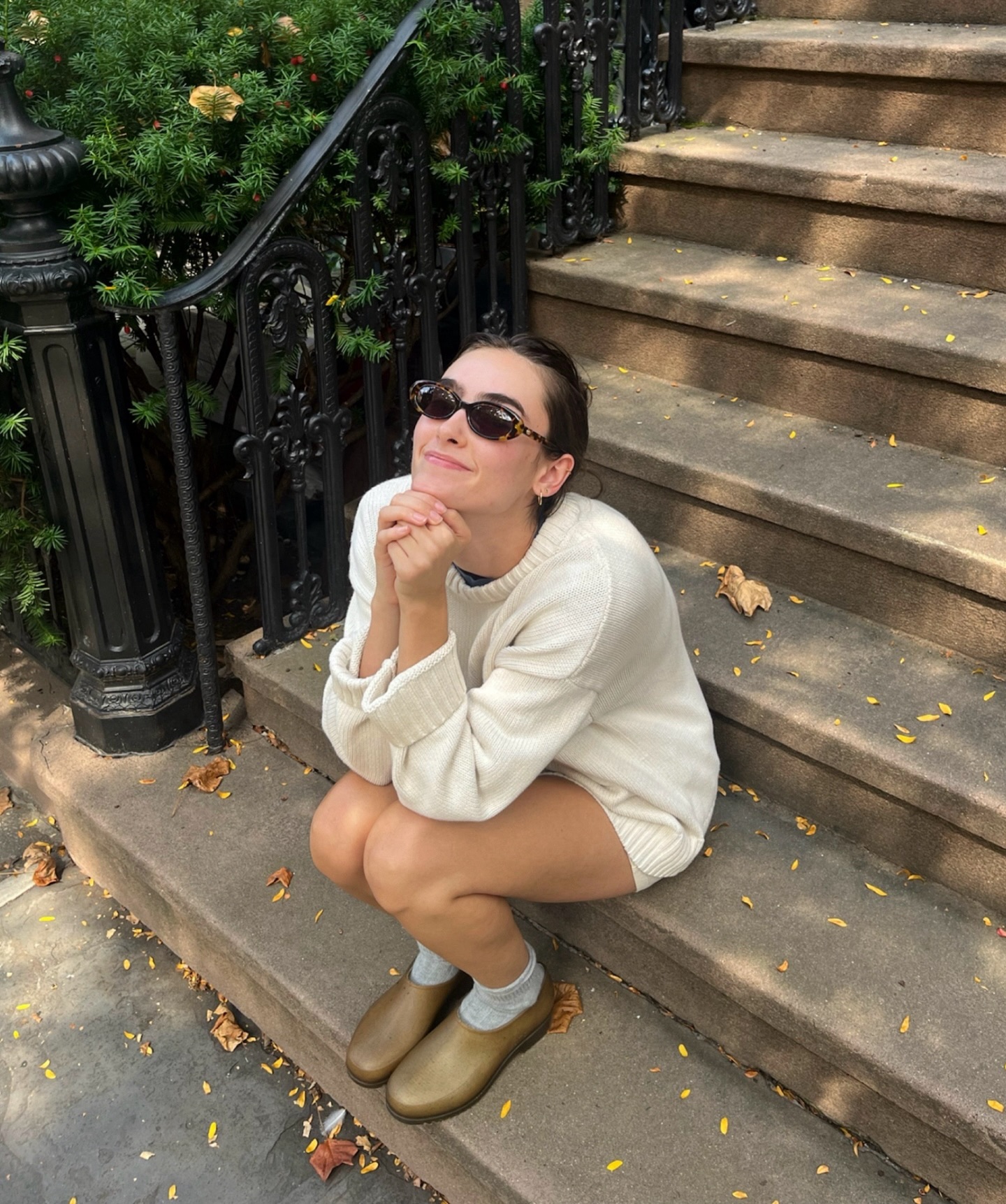 Georgia Weeks wearing Plasticana brown garden clogs, grey socks, a white sweater, black sunglasses, and sitting on steps.