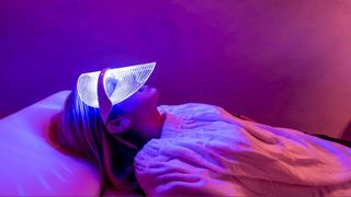 A woman laying on her back wearing an LED face mask emitting red light