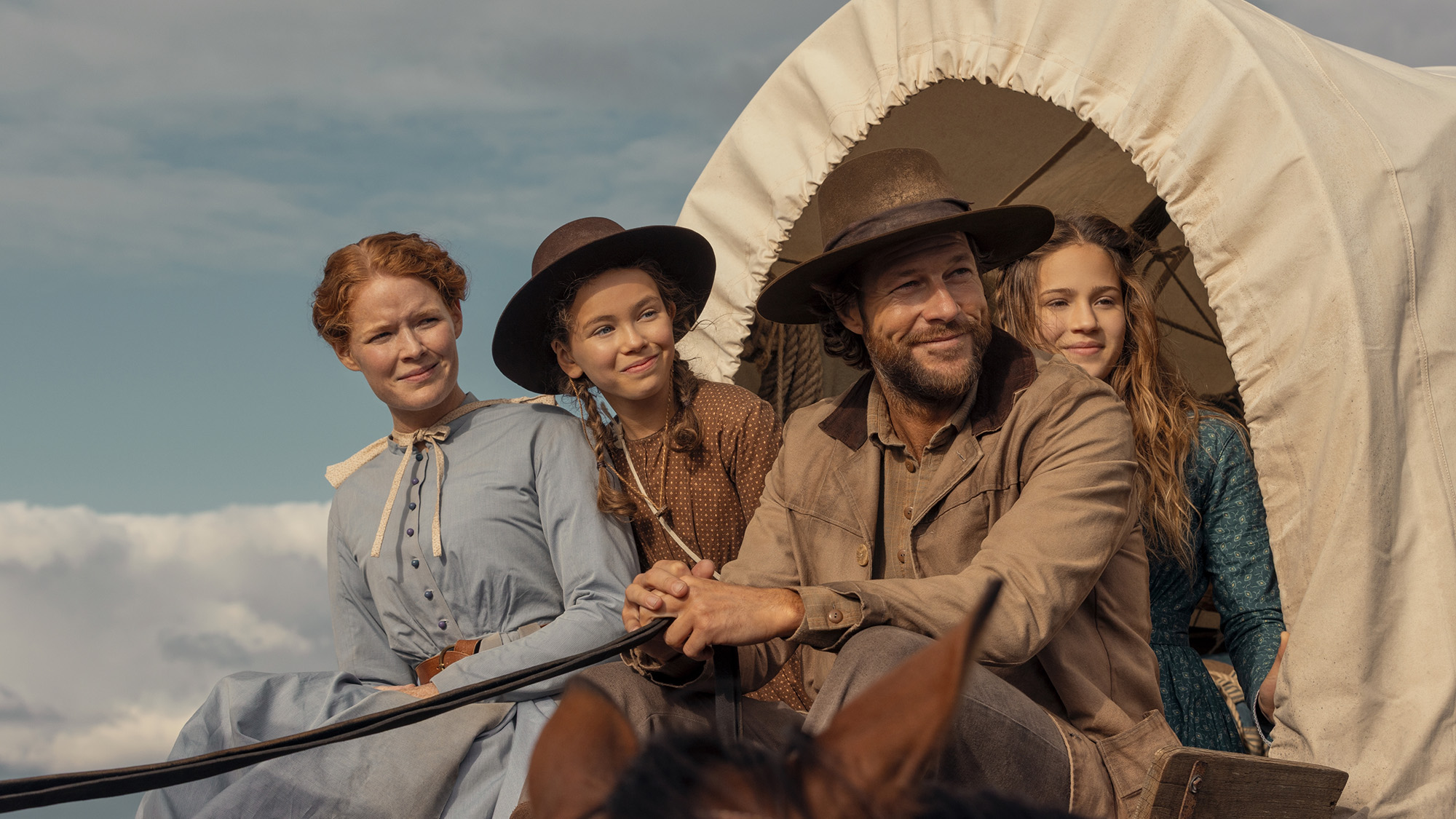 The cast of Little house on the Prairie in long-sleeved cotton period costumes sitting in a covered wagon.