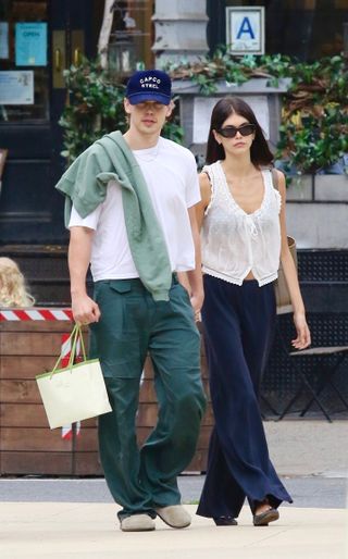 Austin Butler wearing a navy blue hat, white T-shirt, and jeans, with girlfriend Kaia Gerber, wearing a pretty white embroidered top with navy jersey pants, brown ballet flats, and sunglasses walking in NYC.