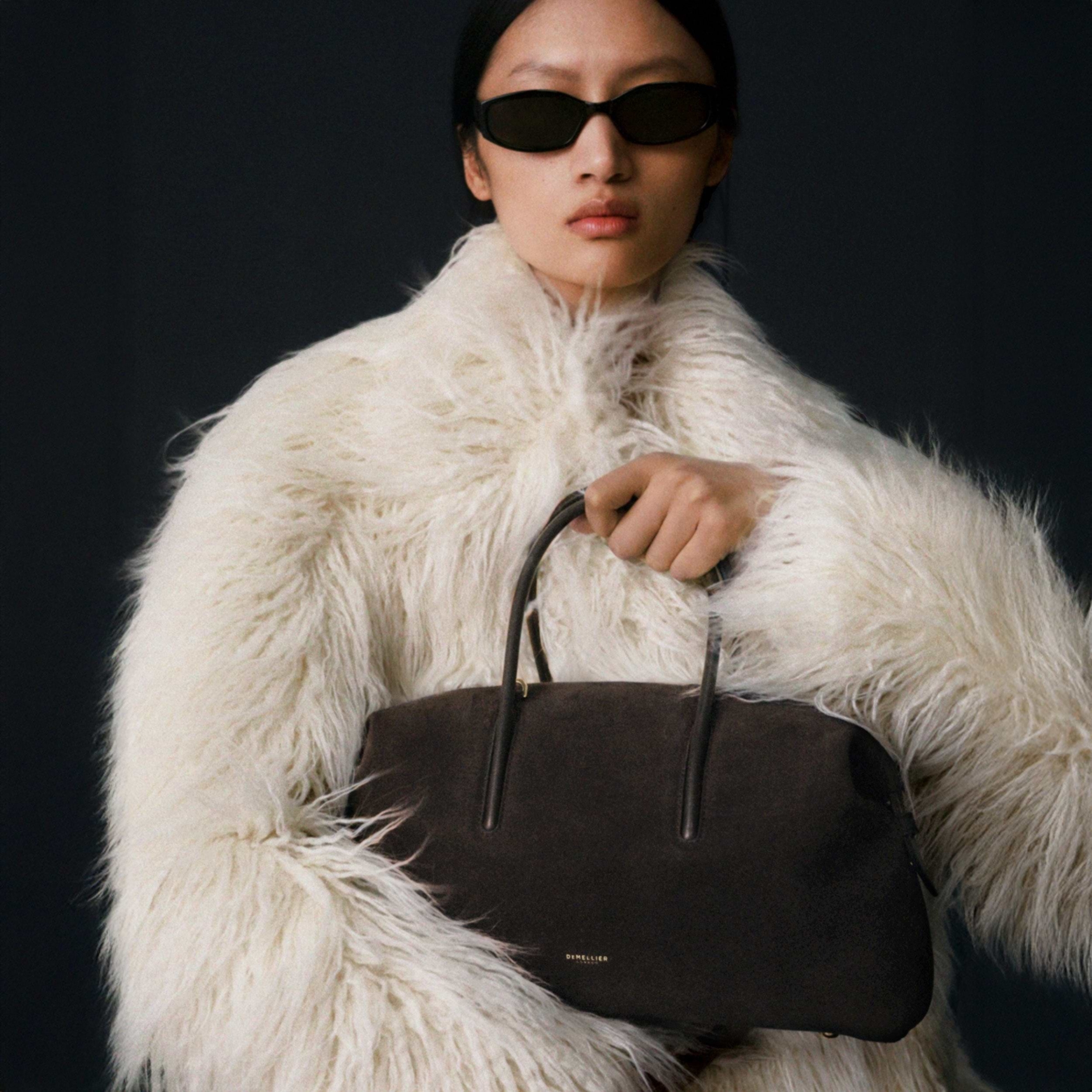 A model wearing a fluffy white coat and holding a brown handbag from DeMellier.