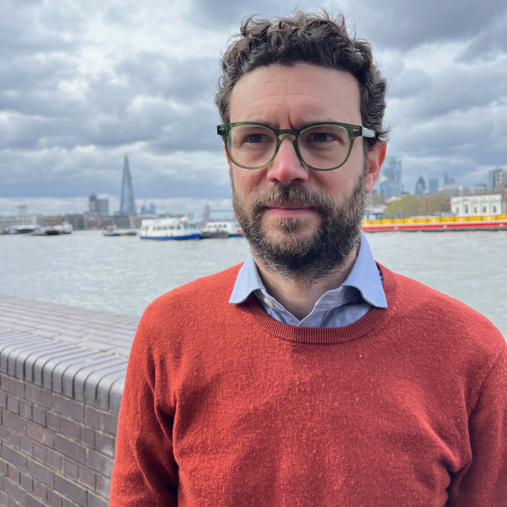 male wearing red jumper, blue shirt with glasses, short hair and beard stood by the River Thames
