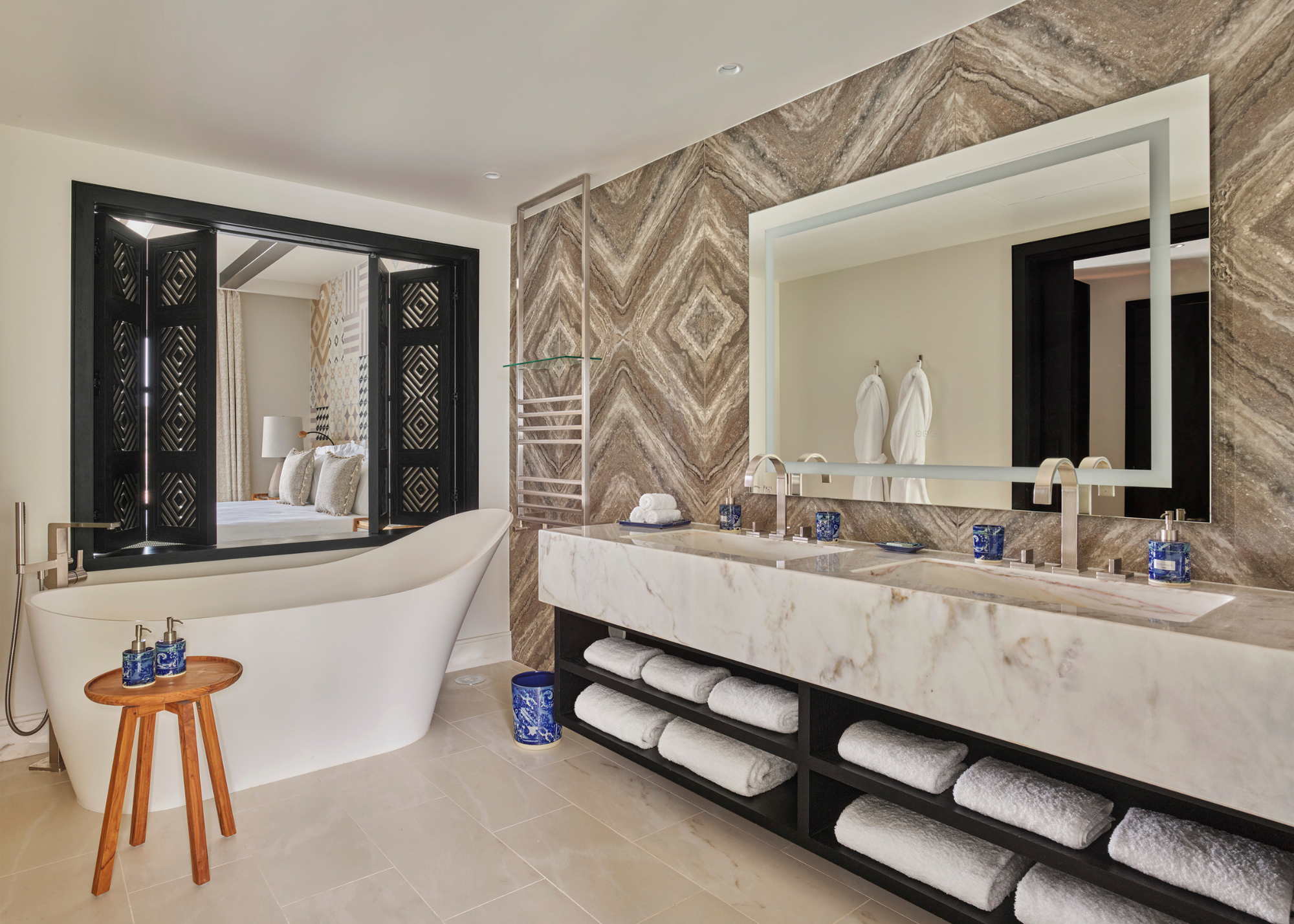 A shot of a bathroom with bookmatched tiles on the wall and a double sink with open shelving at the bottom. There is also a bath tub in the bathroom. 