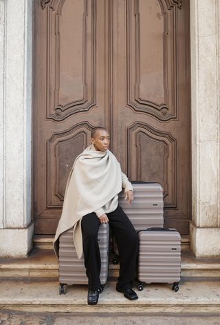 A winter travel outfit is shown in a photo of a woman sitting on top of tan luggage in a doorway, waiting for a taxi while wearing a tan cashmere wrap scarf over a matching sweater styled with black trousers and black loafers