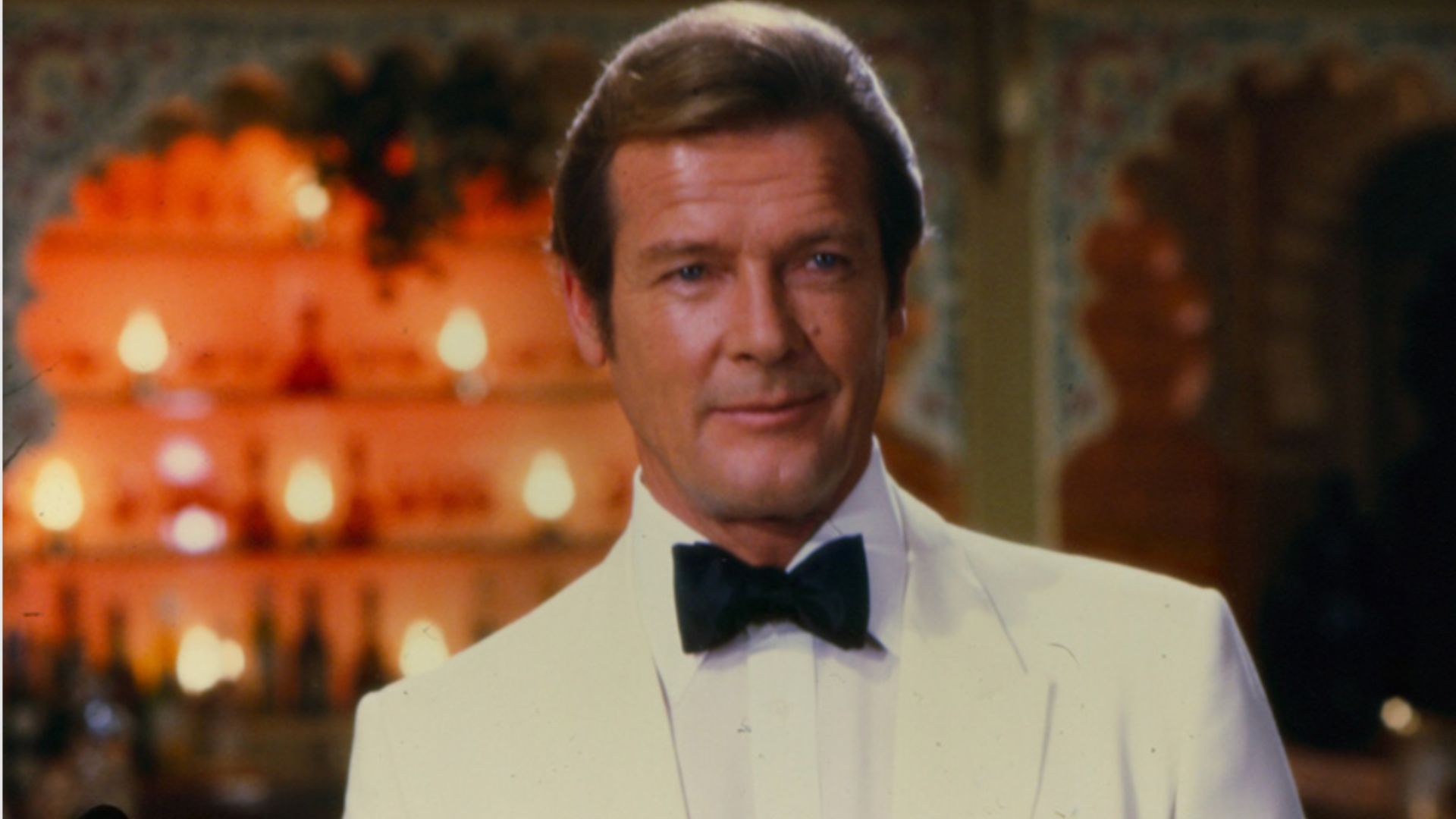 an image of Roger Moore in the bond film Octopussy. He is on screen wearing a white tux with a black bow tie in a bar