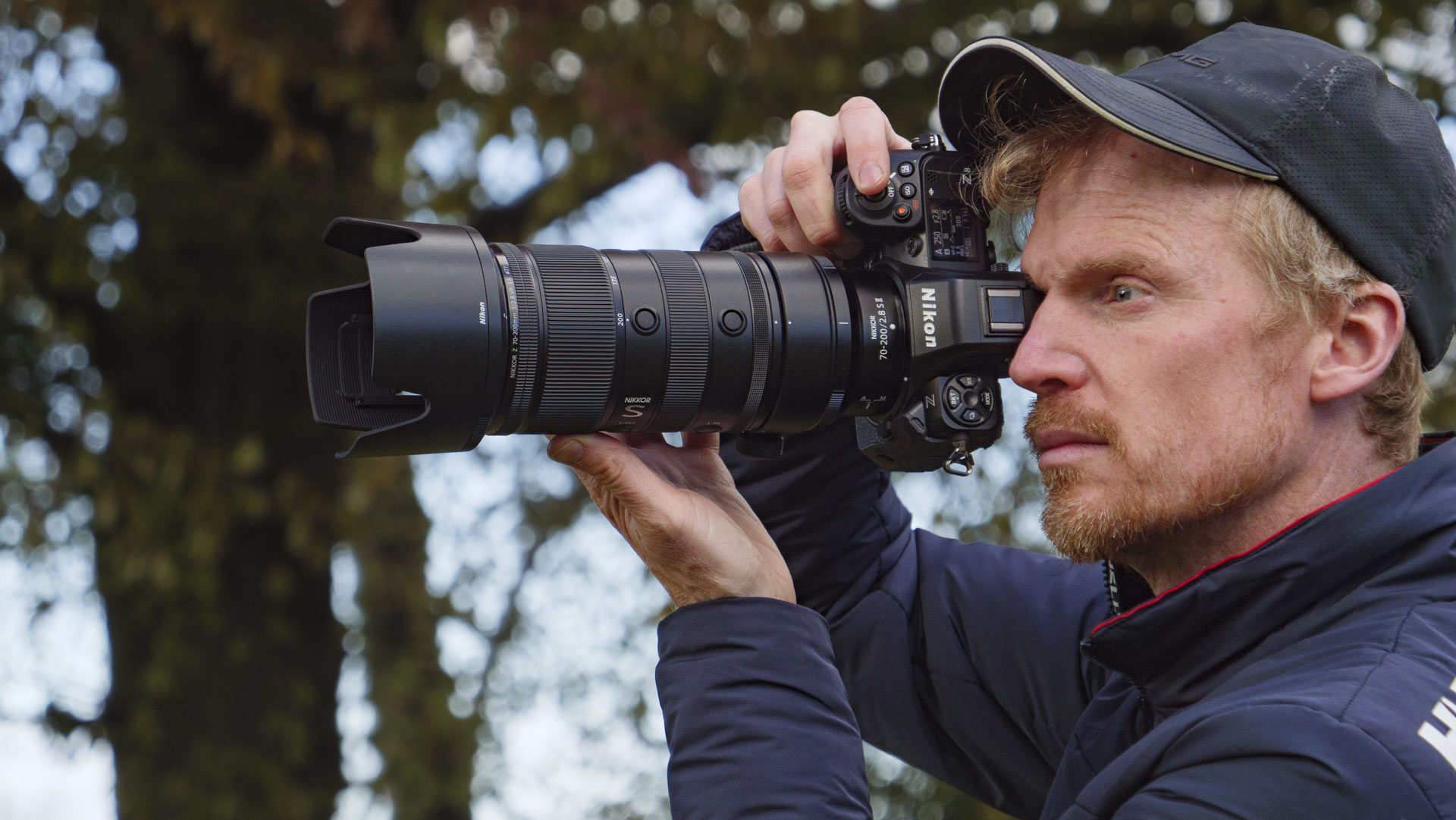 Man holding the Nikon Z8 camera up to his eye and the Nikon Z 70-200mm f/2.8 VR S II lens is attached, there are trees in the background