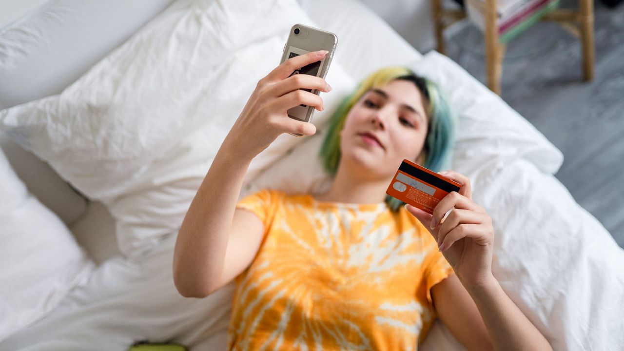 Young woman with colored hair lying in bed looking at her phone and a credit card