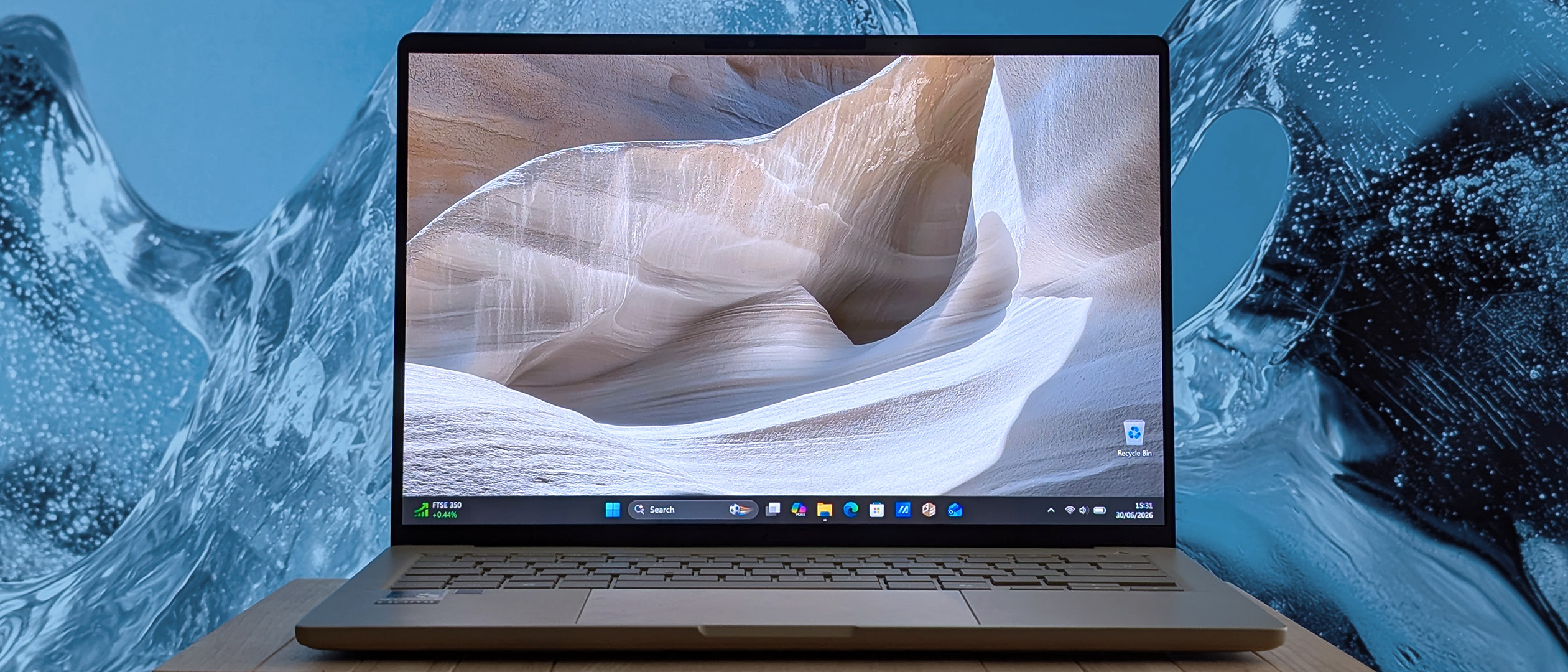 A Zenbook A14 sits on a wooden surface, its display showing a sandstone canyon wallpaper in soft beige and white tones. The backdrop behind the laptop features a cool blue, ice‑like texture.