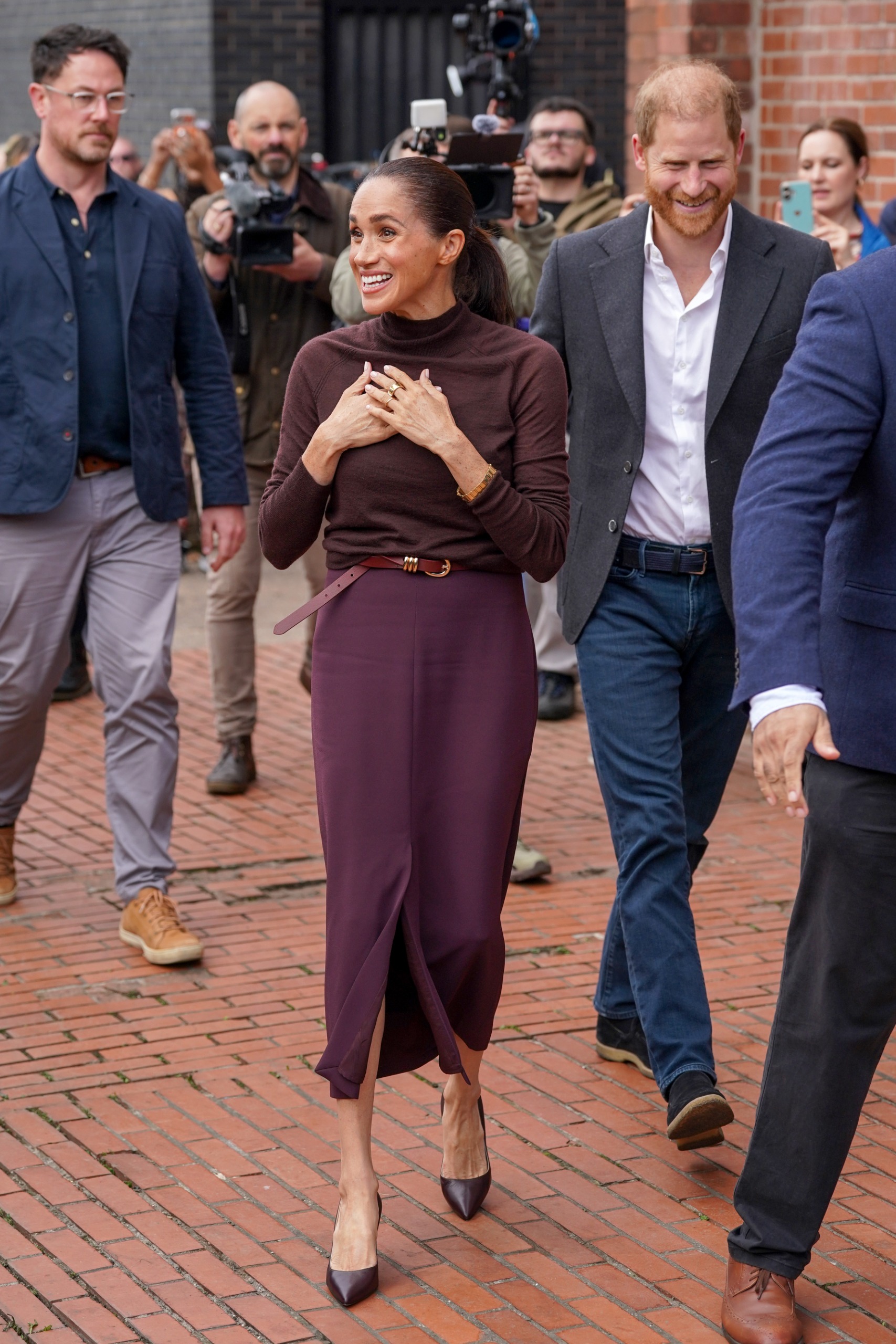 Meghan Markle wearing a maroon skirt and turtleneck putting her hands on her chest as she walks in front of Prince Harry
