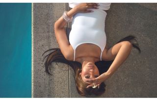 PinkPantheress laying by the pool wearing a white tank top with matching pants and clear chunky jewelry.