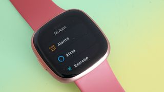 Fitbit Versa 4 med rosa armband mot en grön bakgrund.