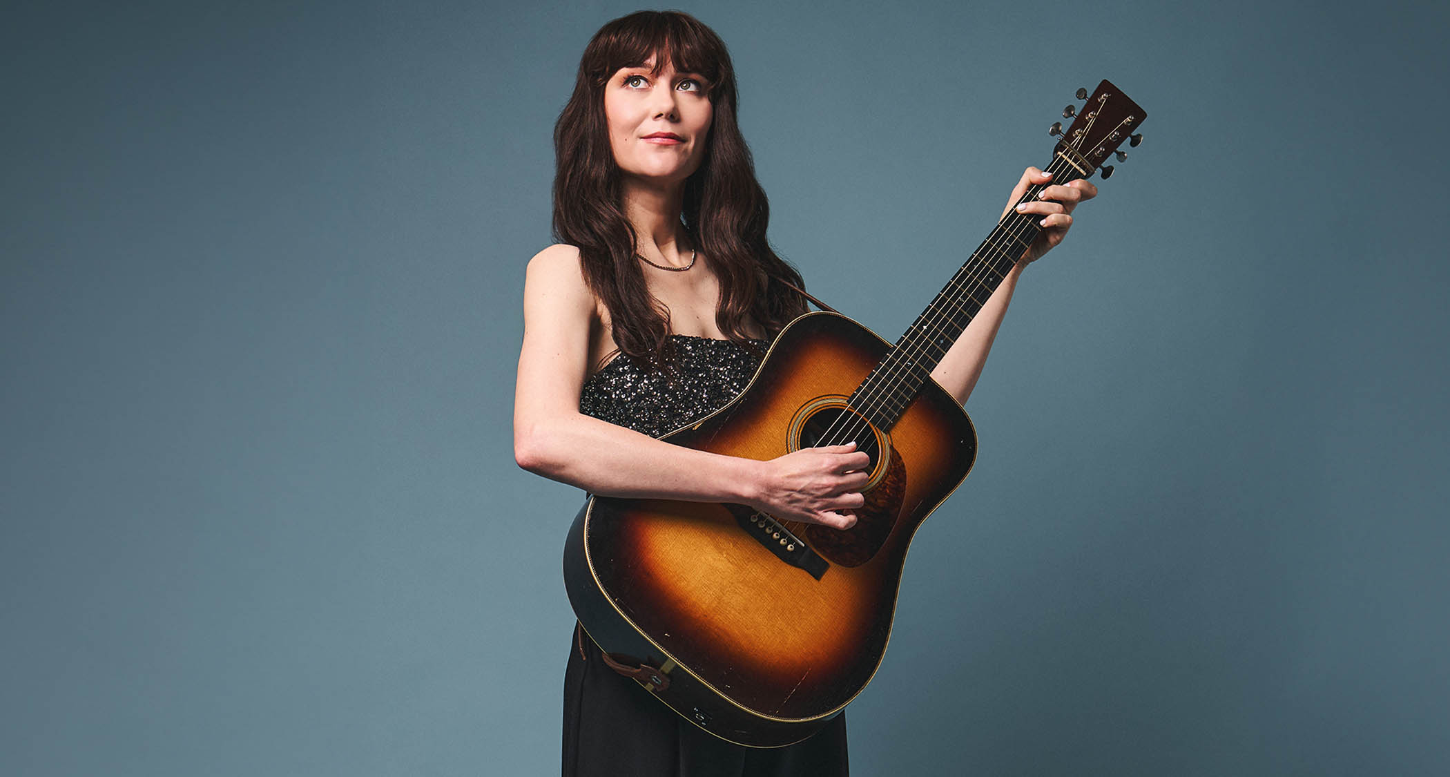 Molly Tuttle photographed with her custom Martin acoustic guitar against a blue-gray background.