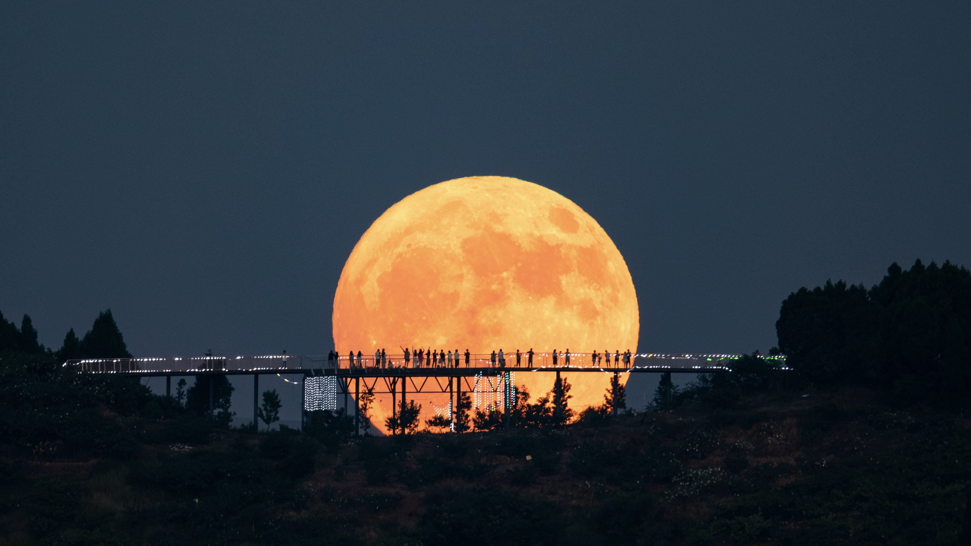 Una gran luna brilla a lo lejos y en primer plano hay una larga plataforma de observación en forma de puente sobre la que se encuentra la gente. Sus siluetas destacan en marcado contraste en la superficie de la luna.