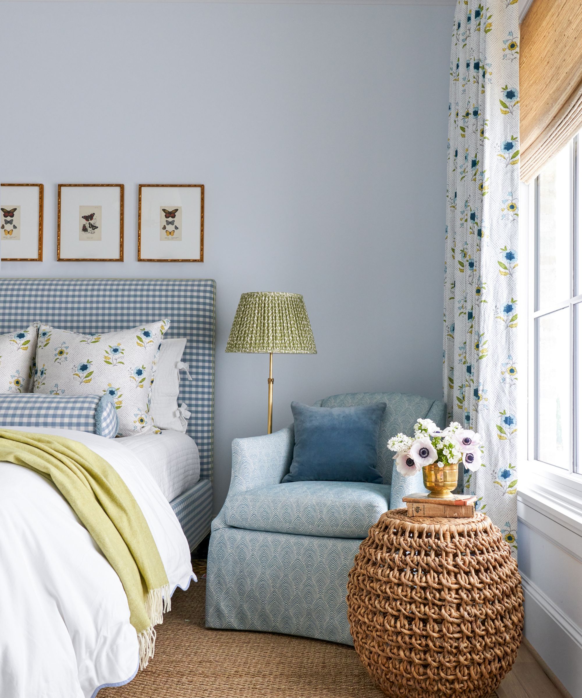 Bedroom with pale blue painted walls, floral curtains and a bamboo blind, a jute rug, a blue armchair in the corner with a floor lamp and a wicker side table, and a blue gingham upholstered bed.