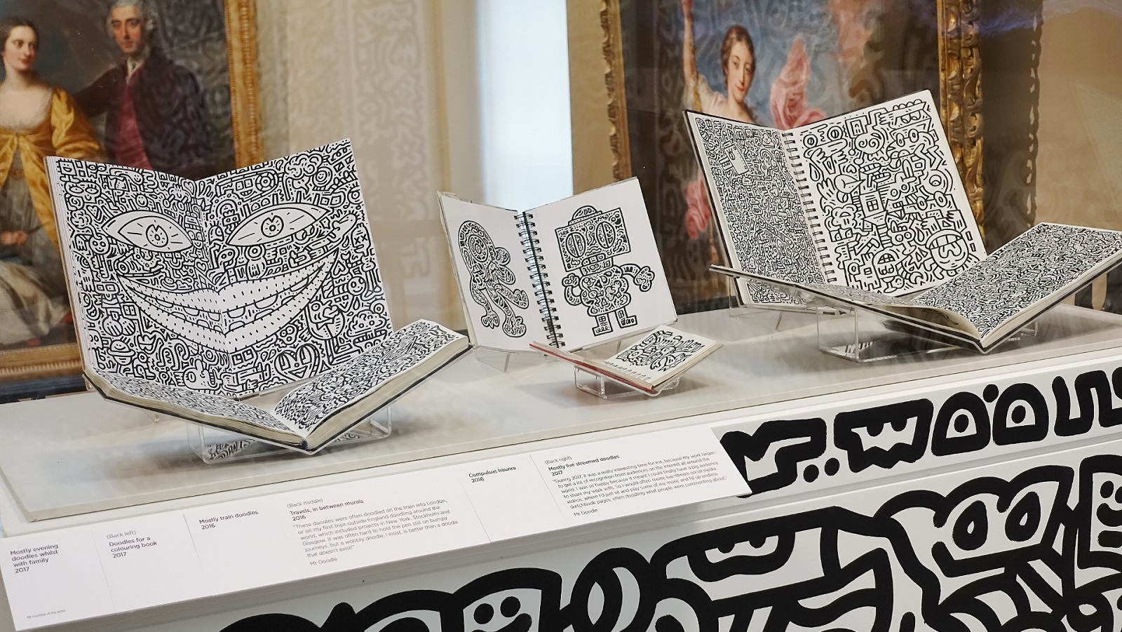 A series of sketchbooks filled with black and white doodle art by Mr Doodle, in a glass display case. Behind them are classical pil paintings, in contrast to the modern doodling.