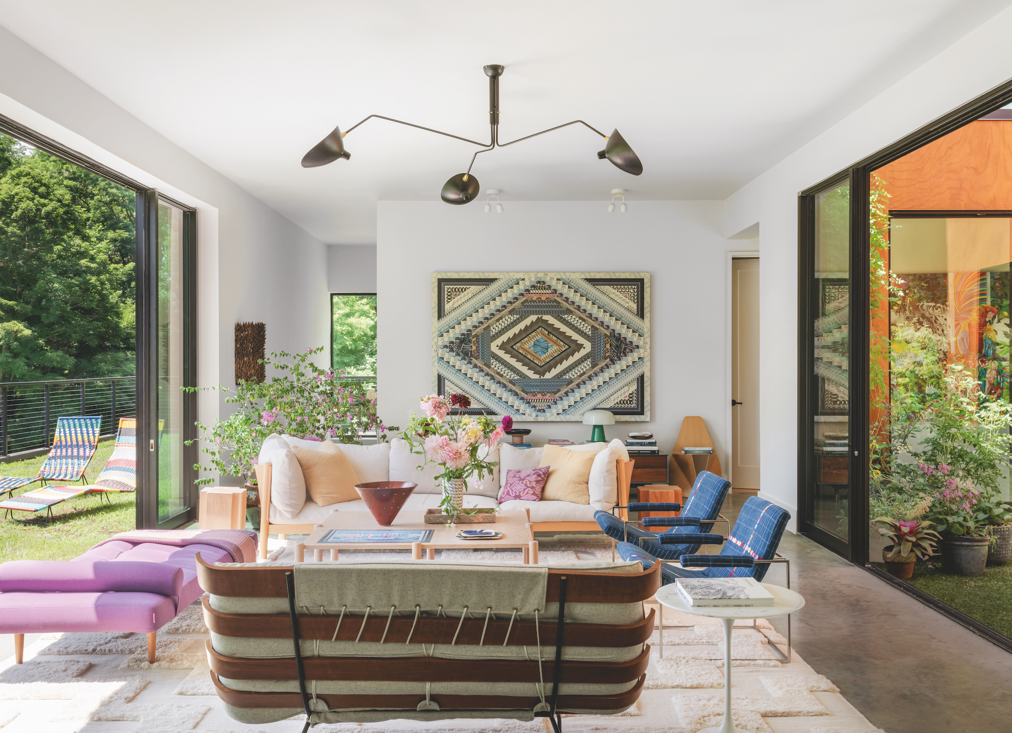 A white, modern living room with sliding doors to the outdoor space on the left and large windows on the right. There is a modern, black chandelier hung above the seating area of miss-matched sofas and armchairs. with an artwork on the wall behind.