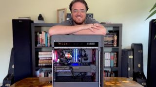 A smiling man leans over a computer case showing colorful internal components, with a bookshelf filled with books in the background.