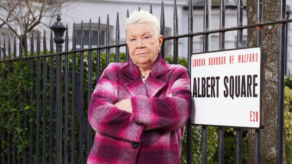 Mo Harris with her arms crossed posing by the Albert Square sign and wearing a pink checked coat for EastEnders 