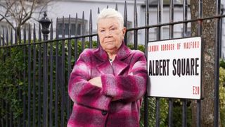Mo Harris with her arms crossed posing by the Albert Square sign and wearing a pink checked coat for EastEnders 