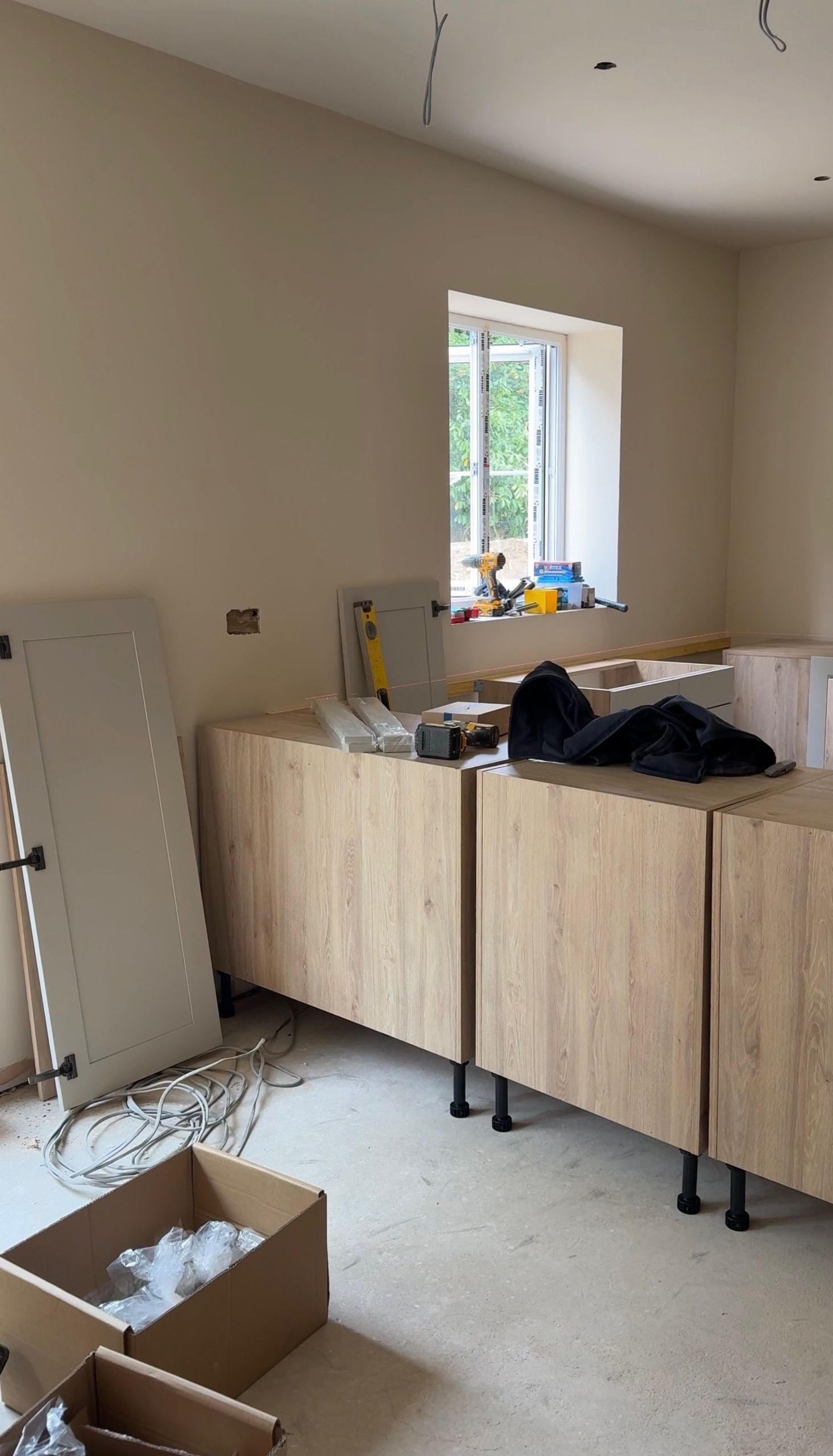 Unfinished room during kitchen installation with light wood lower base cabinets set up along painted walls, tools on top, unmounted cabinet doors, and cardboard boxes on the floor
