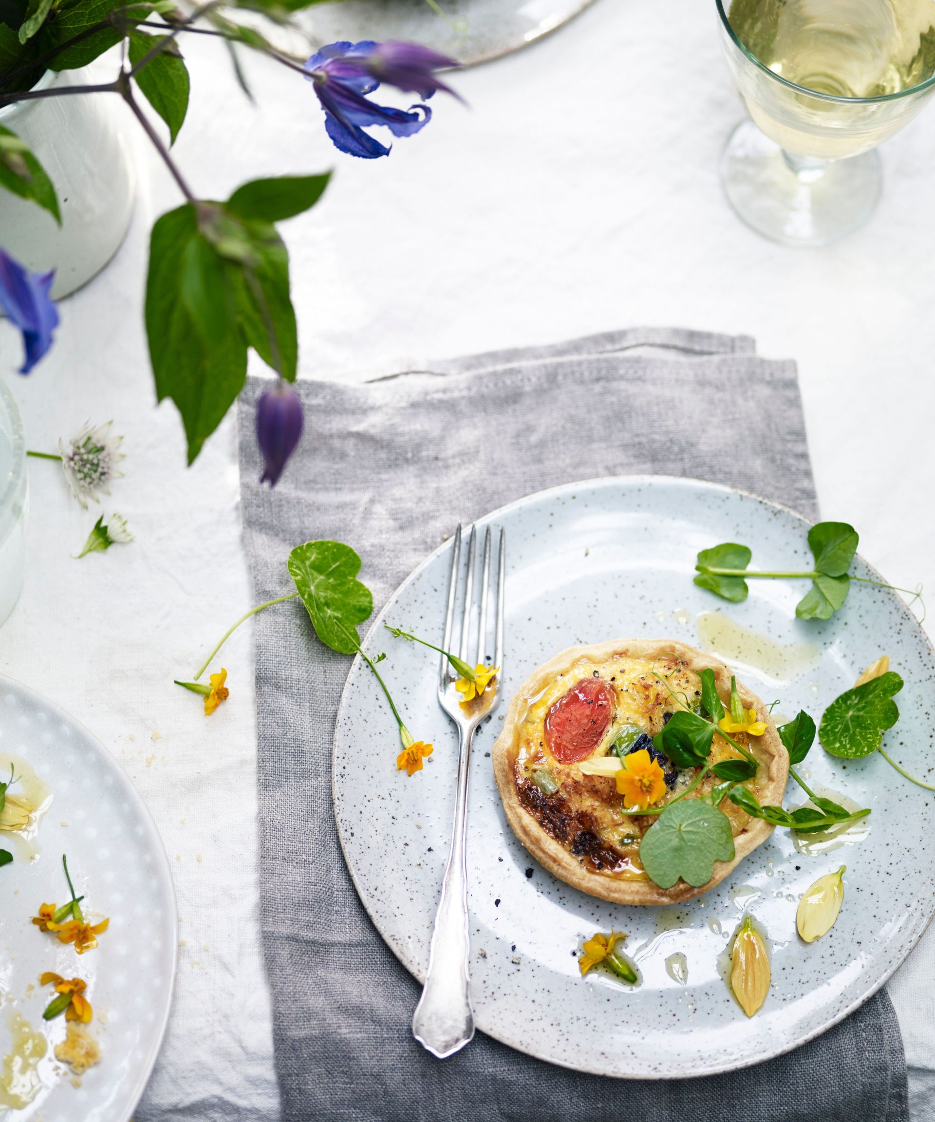 crab tart scattered with edible flowers on a speckled plate on a grey napkin on a white table with a glass of white wine