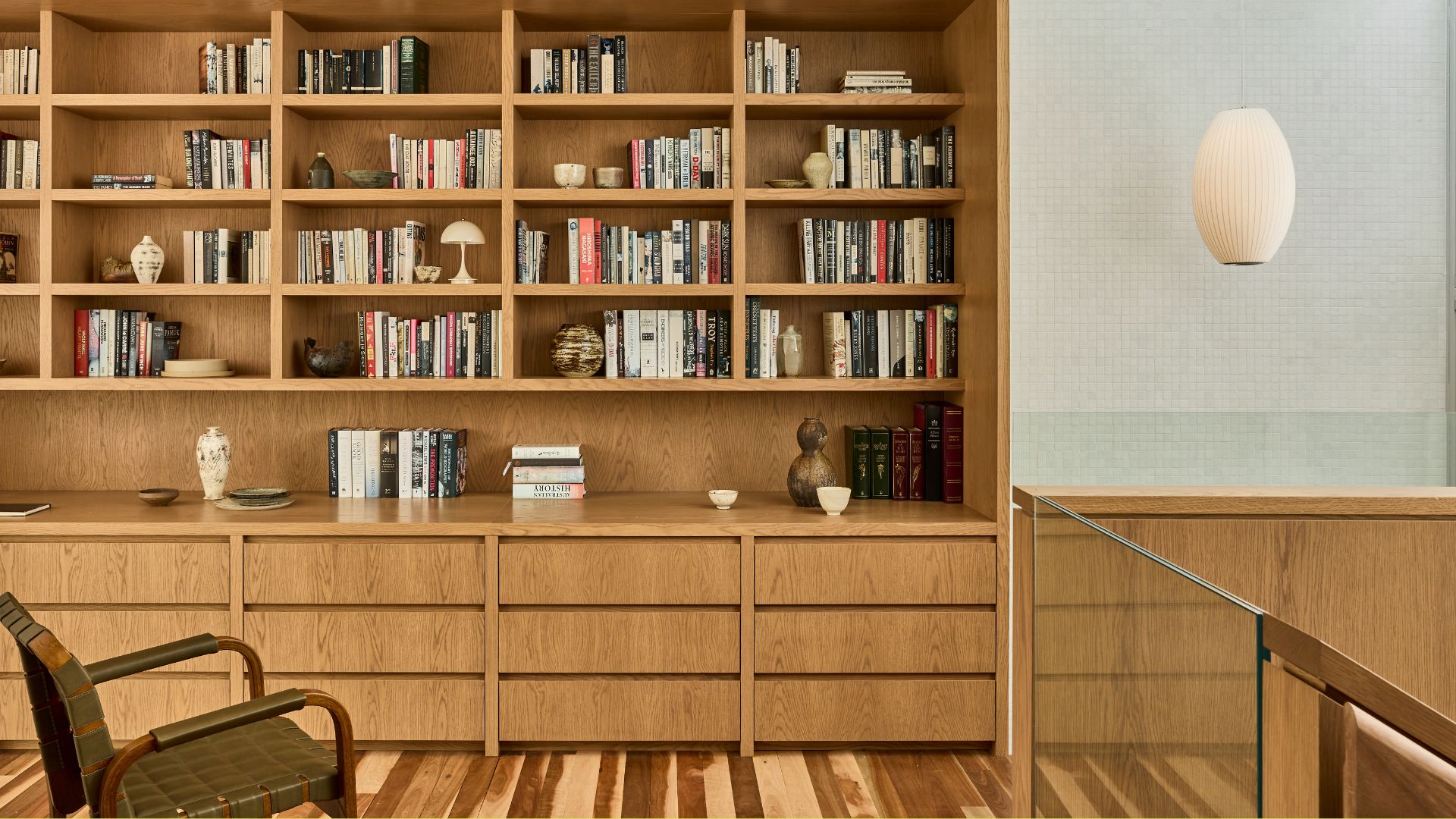 A wood bookshelf wall with colorful books, lamps, ceramics, and a pendant lamp. Also pictured is a green woven chair.