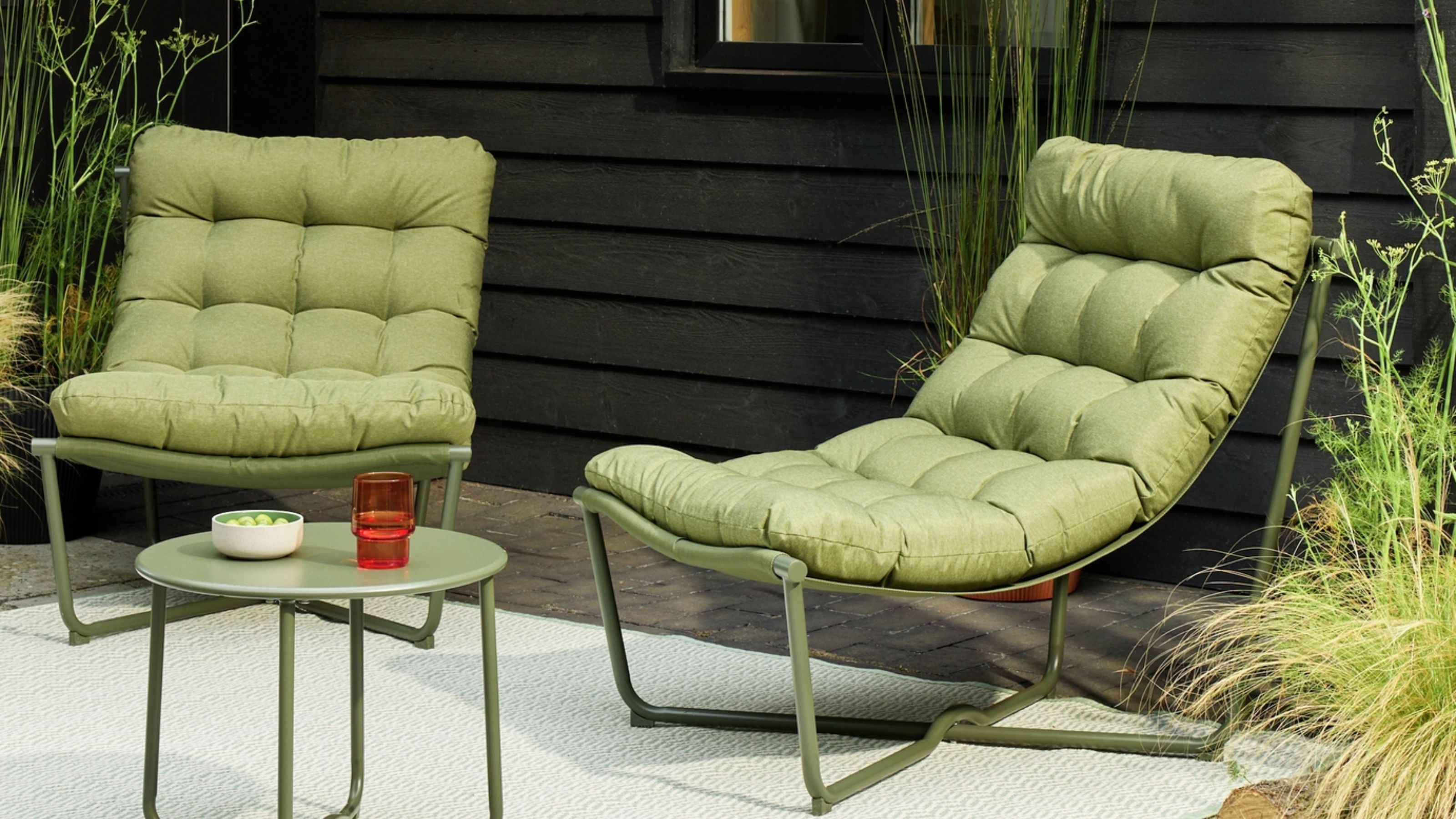 Image of two modern, green outdoor chairs with green cushions positioned around a matching green metal side table. There is a rug underneath them, and the house behind the chairs is dark wood.