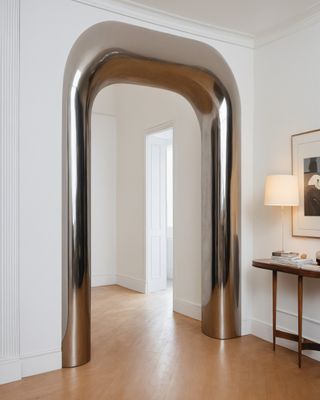 Image of a room with white walls and hardwood floors. The arched doorway is adorned with an oversized, chrome door reveal. There is a wooden console table just inside the room with a lamp on it and a framed picture hanging above.