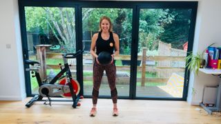 Personal trainer Maddy Biddulph stands holding a medicine ball in her hands in a living room. She wears a vest, leggings and sneakers. Behind her we see a desk, an exercise bike and a large window leading to a grassy outdoor area.