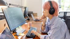 Asian female software developer with pink hair working on computer code on a desktop computer in an open plan office space, with laptop screen open on desk.