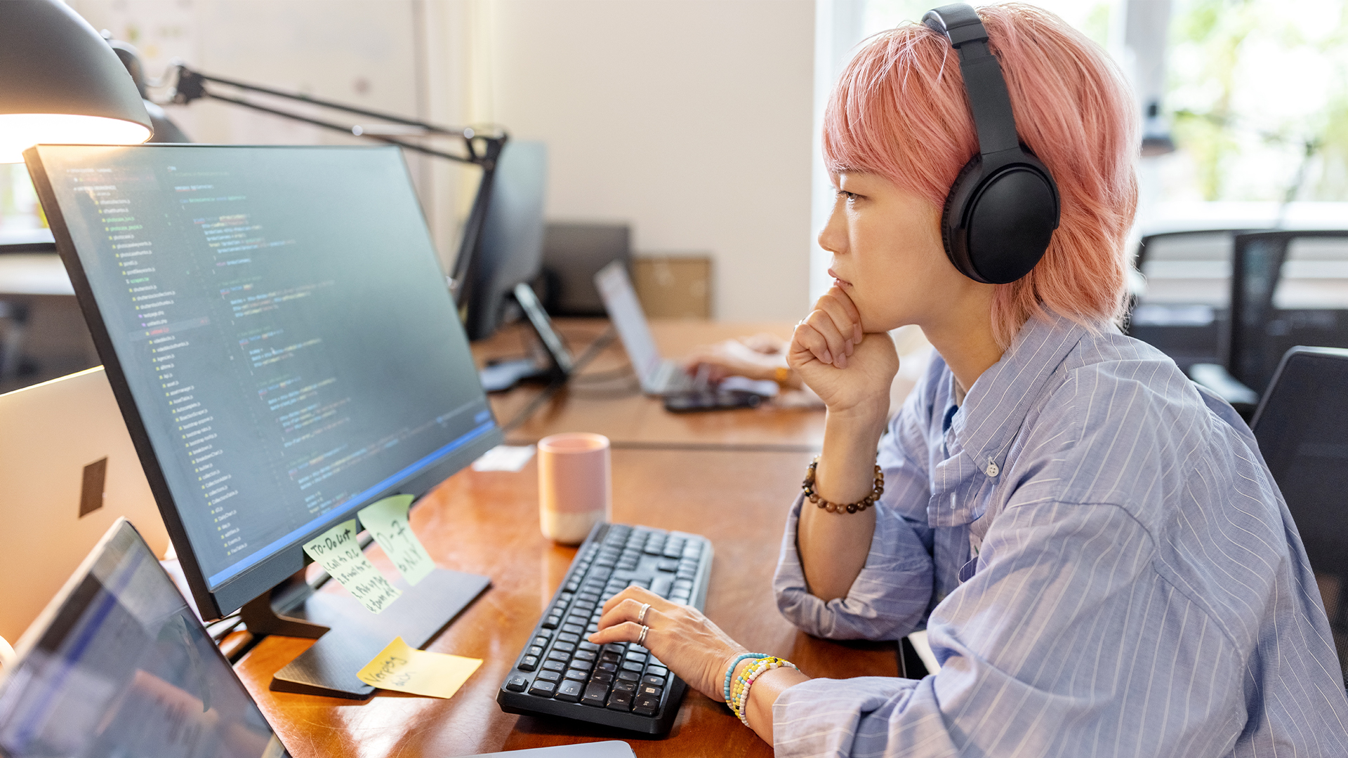 Asian female software developer with pink hair working on computer code on a desktop computer in an open plan office space, with laptop screen open on desk.