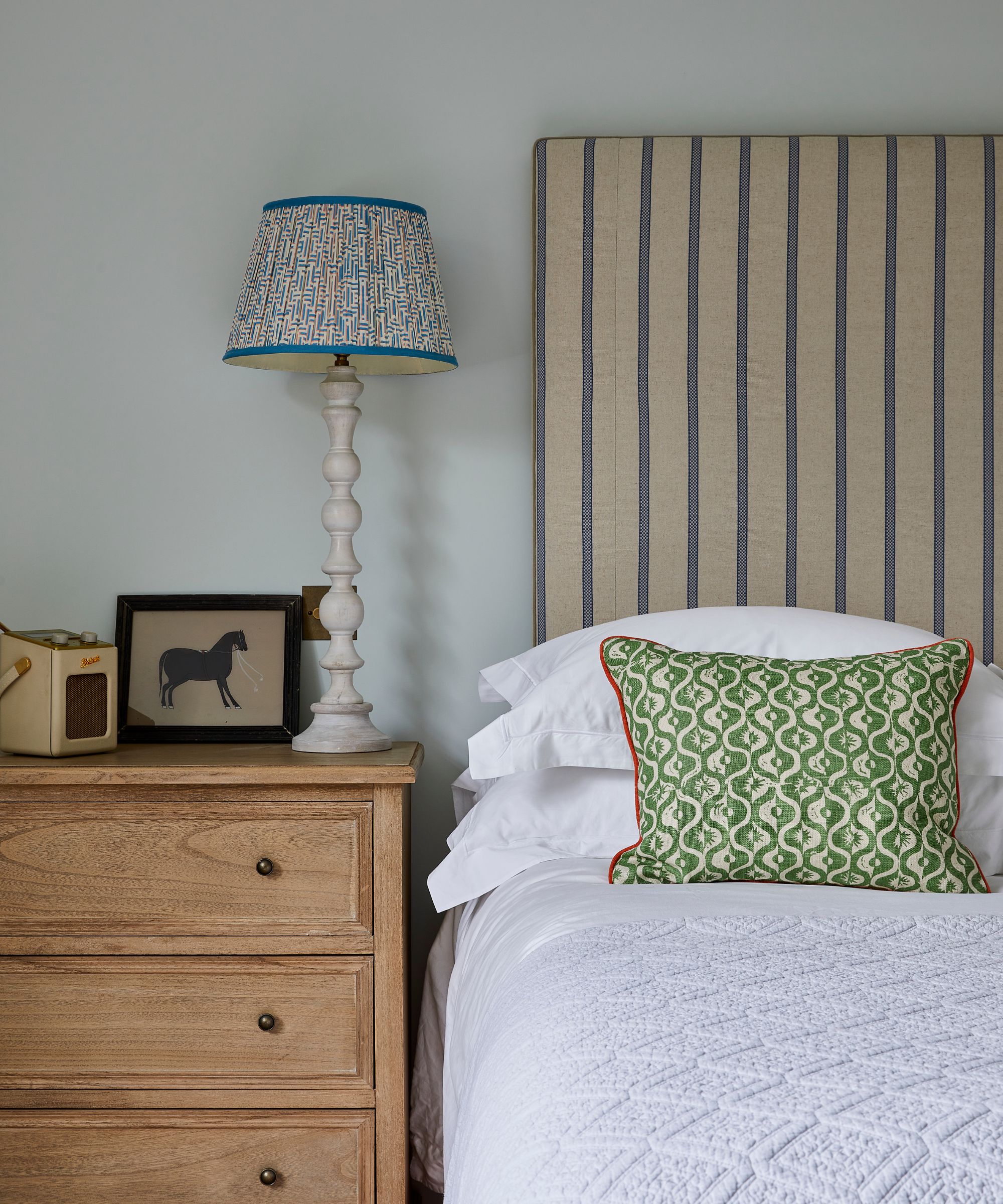 Bedroom painted in Farrow &amp;amp; Ball's Borrowed Light, an upholstered striped headboard, white bedding, a green patterned pillow, and a wooden nightstand with a lamp and an equestrian print on top.