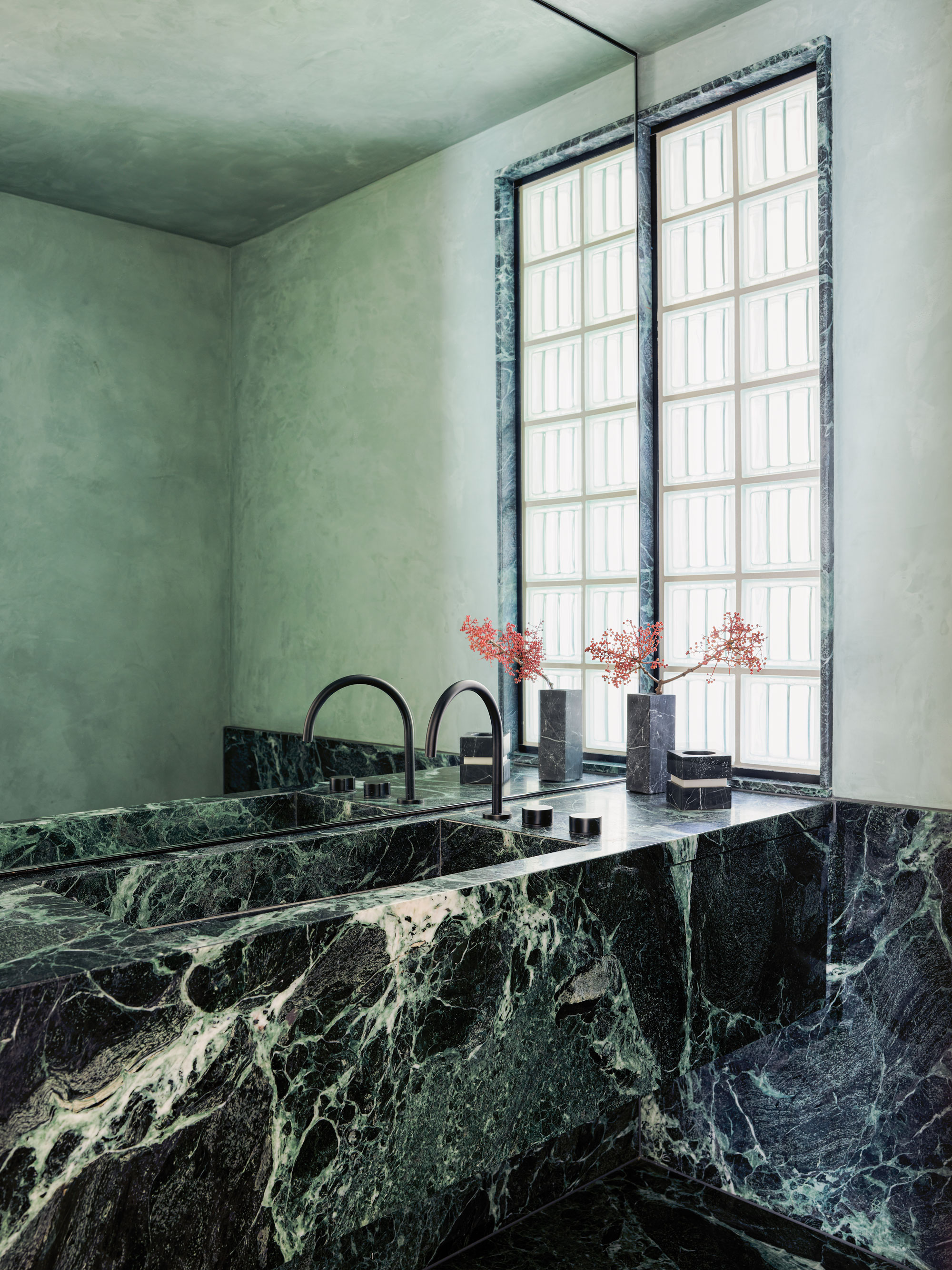 green bathroom with large wall mirror, glass block window, plaster walls in a soft green, and striking green slab sink with vase filled with pink flowers on it