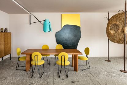 dining room with grey marble flooring, a brown wooden table with six mustard yellow dining chairs, and abstract art including a blue lightshade, wall art and freestanding wooden nest sculpture