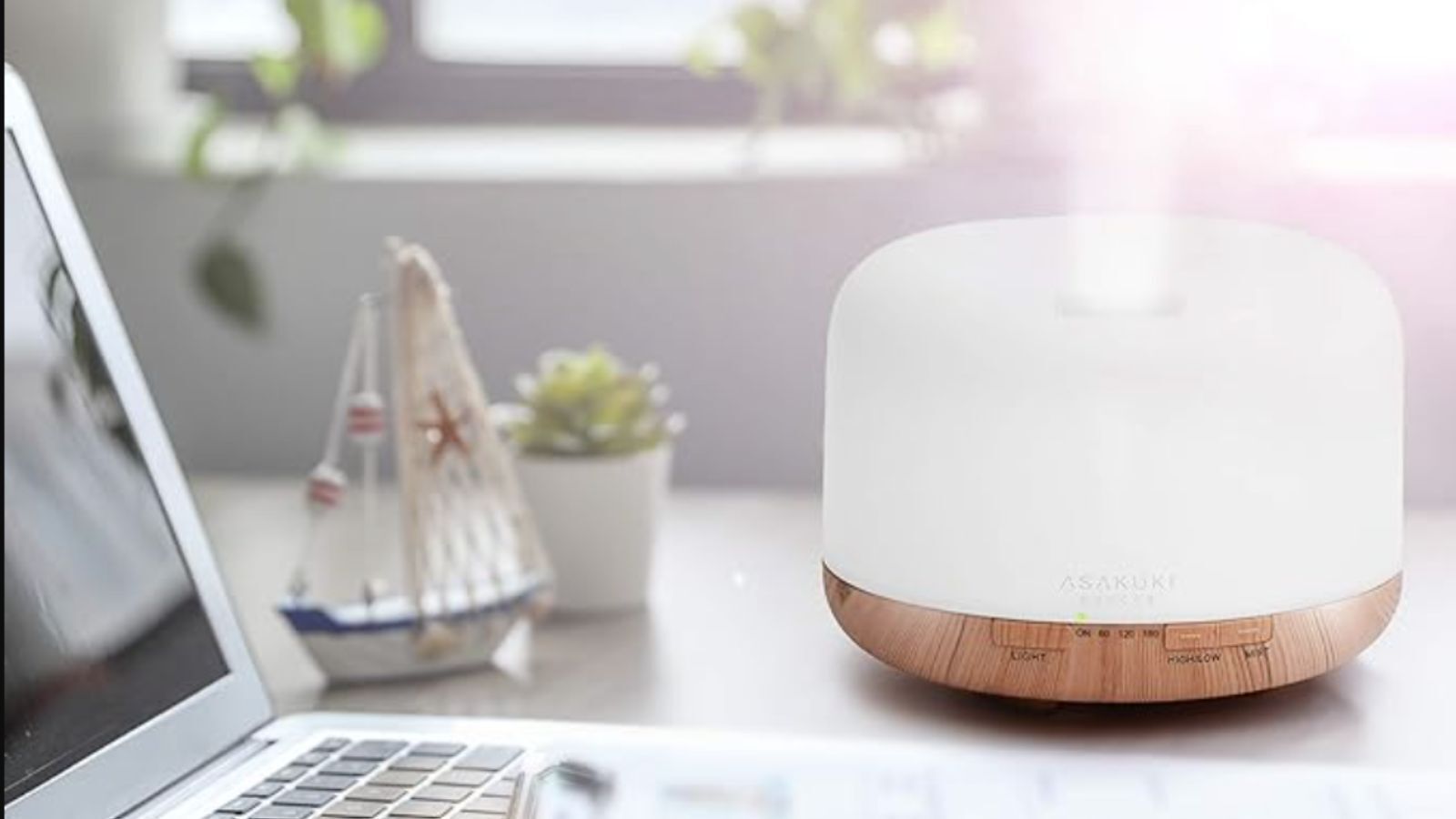 White essential oil diffuser with wood-effect base, sitting on a desk next to a laptop and a small toy boat
