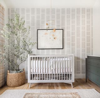 neutral nursery with grey printed wallpaper on focal wall, white crib, plant in basket, artwork, mobile above crib, dark gray changing area with drawers, wooden floor