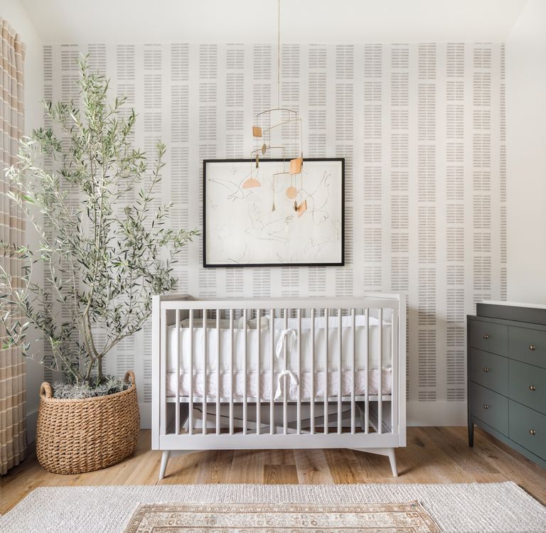 neutral nursery with grey printed wallpaper on focal wall, white crib, plant in basket, artwork, mobile above crib, dark gray changing area with drawers, wooden floor