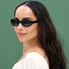 Zoe Kravitz wears a white blouse and cat-eye sunglasses in front of a step and repeat
