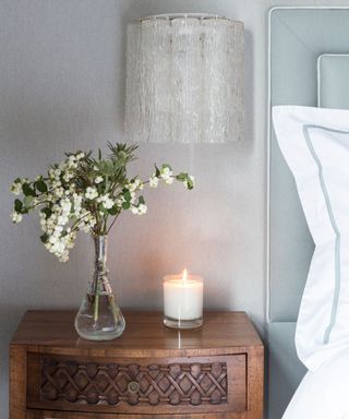 A small candle on a dark wood bedside table beside a glass vase of white flowers