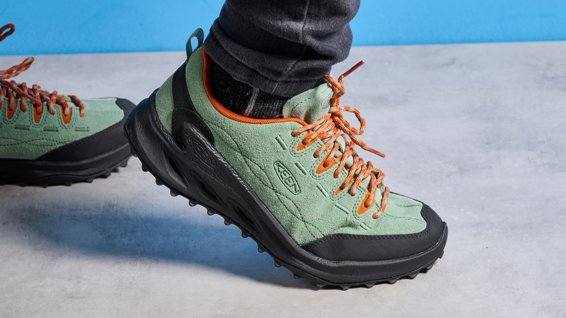 The Keen Jasper zionic hiking shoes in green/gold colorway are photographed against a blue Tom&#039;s Guide background and a stone floor, showing the lug sole and the leather upper with orange laces