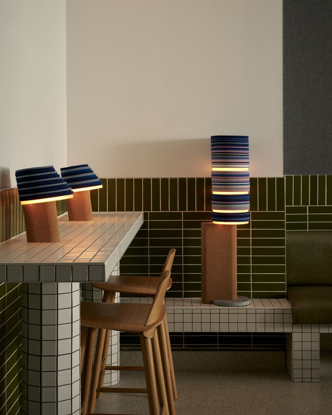 A tiled desk with two wooden chairs, and two striped modern lamps on top. On the far wall sits a taller lamp on a bench seat.