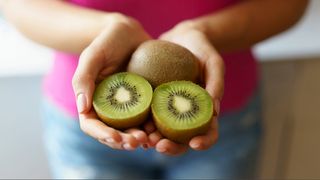 Person holding two kiwifruits in the palm of their hands, one kiwi is cut in half the other is whole.