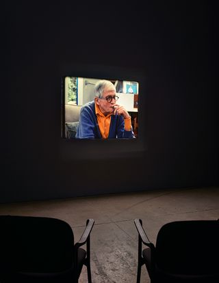 Dark room with two chairs and screen showing David Hockney