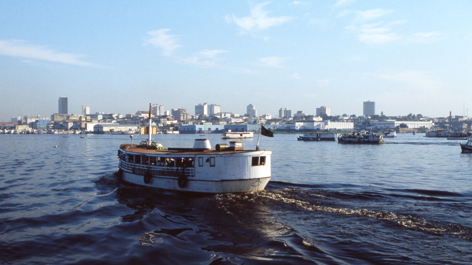 In this image you see a white ferry boat full of tourists as it travels on the Amazon River.  In the background you can see the city skyline of Manaus, Brazil.