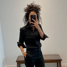 Neelam wears a black button-down shirt, black trousers, and the row signum heels.
