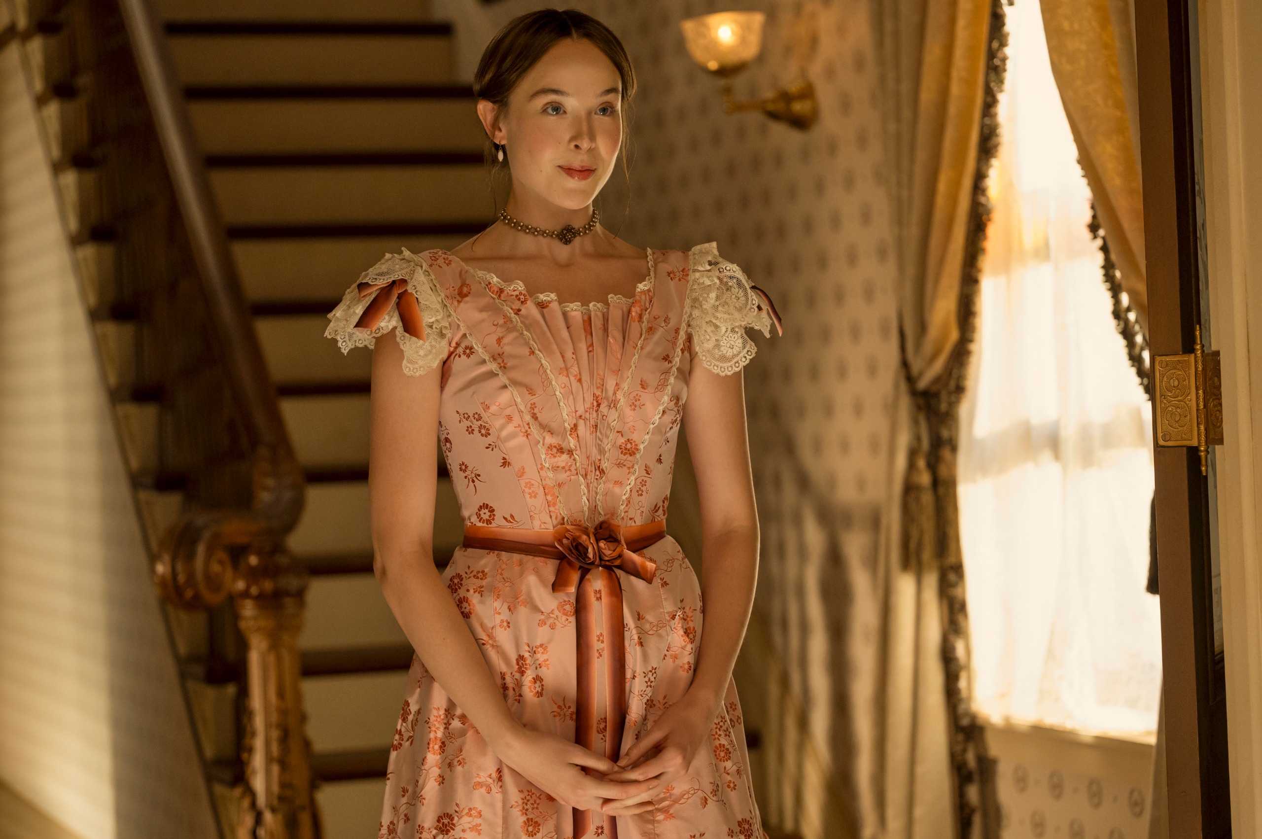 ella beatty wears a pink dress and stands in a victorian era home at the bottom of a staircase in a still from monster the lizzie borden story