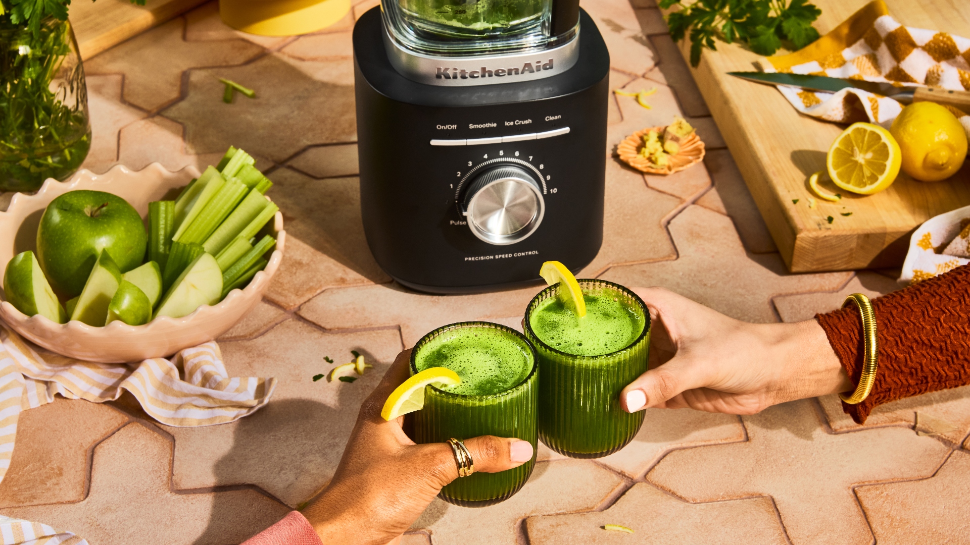 Two hands cheers green drinks with the KitchenAid Pure Power Blender behind